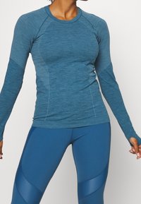 Camiseta deportiva de manga larga en tela azul jaspeado con acentos texturizados, con mangas raglán y un diseño ajustado, combinada con leggings azules.