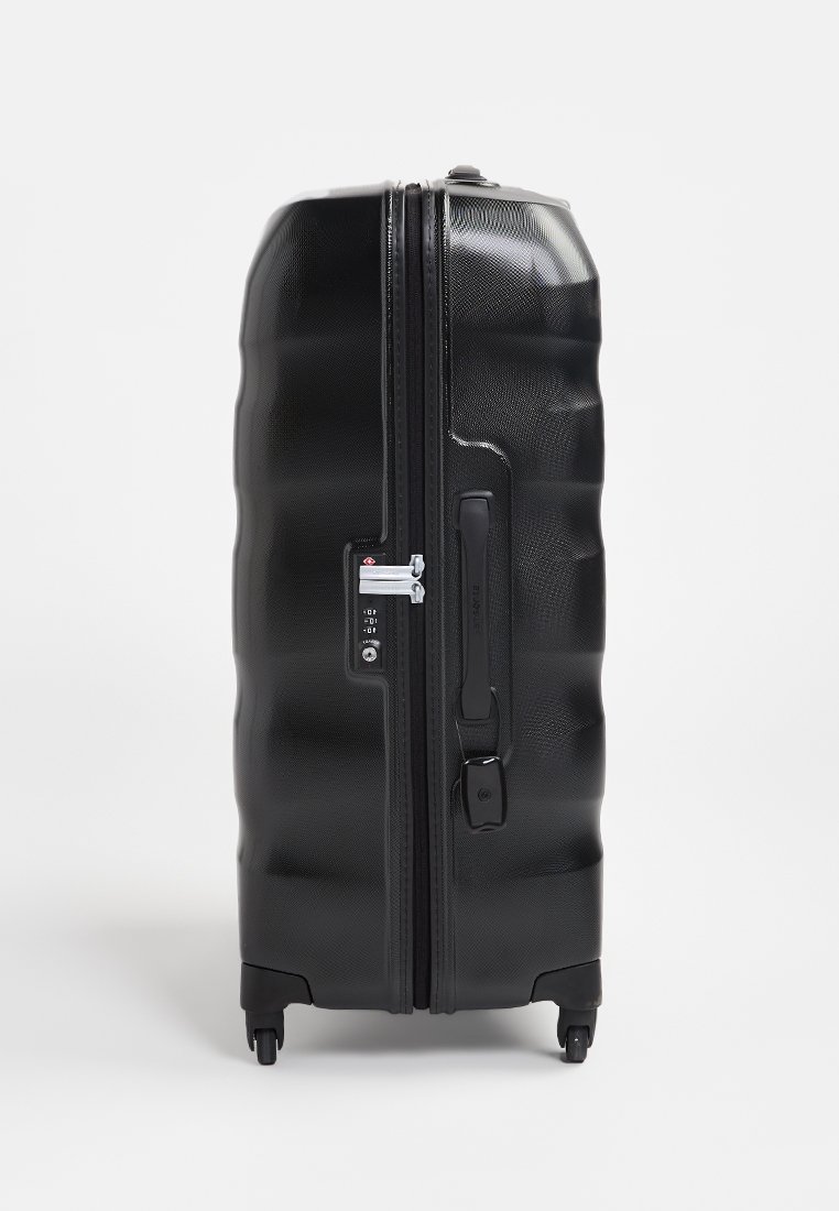 Valise rigide noire avec une surface texturée, rainures verticales, double zip, serrure à combinaison et quatre roues pivotantes pour une mobilité aisée.