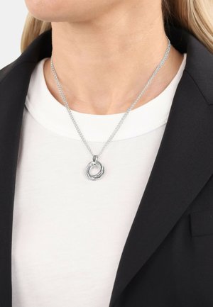Silberner Metalldekorationsanhänger mit ineinander greifenden kreisförmigen Designs, der an einer feinen Kette hängt, getragen über einem weißen Hemd und einem schwarzen Blazer.