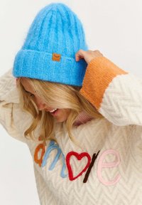 Modrá pletená čiapka s rebrovanou textúrou a koženou nášivkou; krémový sveter s farebnými vyšívanými písmenami a vzorom chevron.