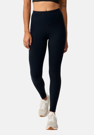 Vrouw die zwarte leggings met hoge taille en witte sportschoenen draagt, staand met één been licht gebogen tegen een effen achtergrond.