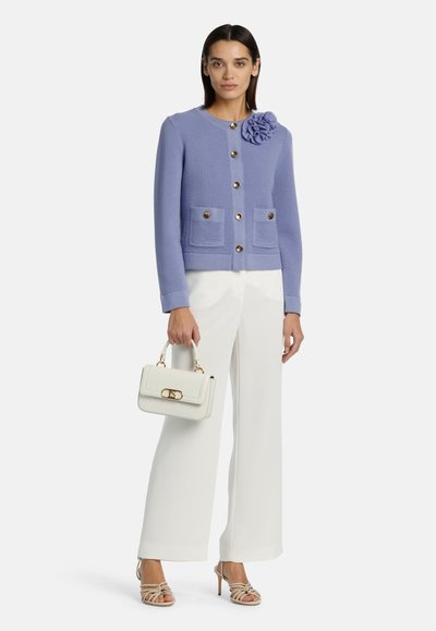 Cárdigan de punto lila con un acento floral, con botones dorados y bolsillos cuadrados. Combinado con pantalones blancos y un bolso crema.