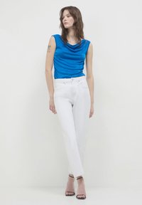 Donna in piedi su uno sfondo bianco, vestita con un top senza maniche blu, jeans bianchi a vita alta, tacchi neri con cinturini, con un tatuaggio visibile sul braccio.