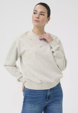 Vrouw draagt een lichtbeige sweatshirt met decoratieve strassstenen aan de bovenkant voorkant, gecombineerd met blauwe spijkerbroek, poseert met één hand bij de kraag.