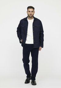 Marineblaue Jeansjacke mit zwei Brusttaschen und braunen Knöpfen, über einem weißen Strickpullover und einer dunklen marineblauen Cordhose. Schwarze Schuhe.