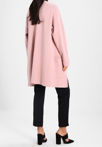 Vaaleanpunainen ylisuuri neuletakki, jossa on rento istuvuus, pitkät hihat ja sivutaskut, yhdistettynä mustiin housuihin ja avokkaisiin.