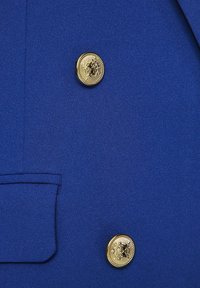 Mėlynas švarkas su lygia medžiaga, dvigubos sagos dizainu ir auksinėmis sagutėmis su emblemos detale. Į komplektą įeina diskretiška kišenė kairėje pusėje.