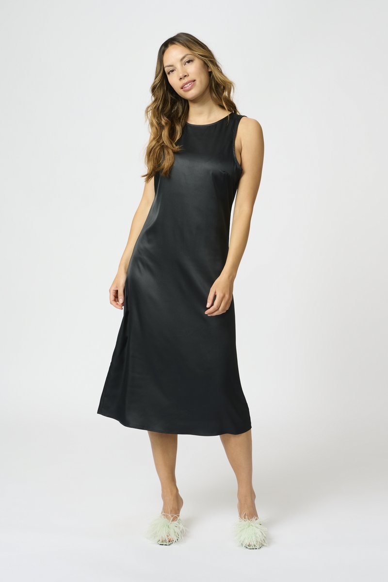 Sieviete stāv slepenā melnā midi kleitā bez piedurknēm ar apaļu dekoltē un valkā baltas spalvu augstpapēžu sandales.