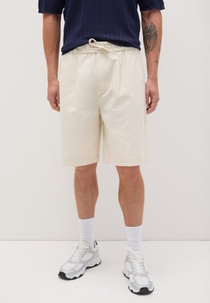 Crèmekleurige shorts met een relaxte pasvorm, plooien aan de voorkant en een elastische tailleband. Gecombineerd met een marineblauw gebreid shirt en sneakers.