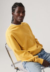 Yellow cotton sweatshirt with a crew neckline, long sleeves, and ribbed cuffs, paired with light blue jeans. Features a subtle logo detail.