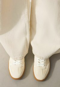 Sneakers lavet af letvægtigt crèmefarvet materiale, med perforerede detaljer, hvide snørebånd og en kontrasterende brun gummisål.