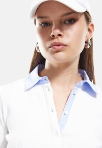 Weißes geripptes Poloshirt mit einem hellblauen gestreiften Kragen, ausgestattet mit Knopfverschluss und einem detaillierten Halsausschnitt. Das Modell trägt eine weiße Kappe und Creolen.