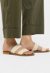 Beige to-straps sandaler med flat såle, med en glatt lærfinish og åpen tå design, sammen med olivengrønne avkorte bukser.