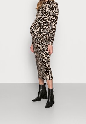 Zwangere vrouw die een getailleerde, lange mouwen midi-jurk met dierenprint draagt en zwarte enkellaarsjes met hakken, staand tegen een effen achtergrond.