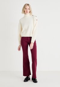 Suéter de punto grueso color crema con un alto cuello y botones decorativos, combinado con pantalones acampanados ajustados color burdeos y mocasines negros.
