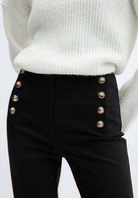 Pantalon noir taille haute avec une texture lisse, muni de boutons décoratifs dorés sur le côté et associé à un pull blanc à côtes.