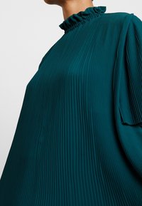 Vestido plisado en color azul verdoso con un escote alto con volantes y mangas anchas. Hecho de una tela texturizada que resalta los plisados verticales y el drapeado suave.