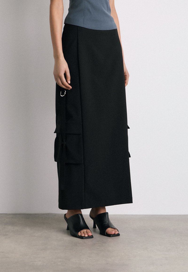 Jupe midi noire avec poches latérales, détails en métal et coupe droite, assortie à des mules noires à bout ouvert sur un fond neutre.