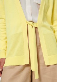 Geel vest met textuurmesh zakken, open voorkant en striksluiting. Gecombineerd met beige broeken, die het zachte, lichte materiaal benadrukken.