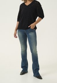 Pull noir en tricot avec un col en V et des manches trois-quarts, associé à un jean évasé bleu et des chaussures noires à bout pointu.