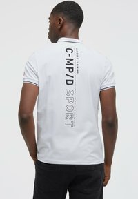 Polo blanc avec col et poignets noirs, présentant un texte noir imprimé verticalement au dos. Tissu doux et design structuré évident.