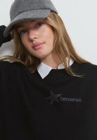 Sweatshirt noir avec une étoile grise et le texte "brownie". En dessous, une chemise blanche à col est visible. Le mannequin porte une casquette en feutre grise.