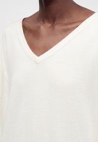 Personne à la peau foncée portant un pull crème à col en V, montrant le cou, la clavicule et la partie supérieure de la poitrine.