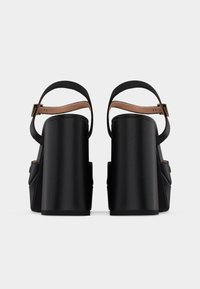 Ζευγάρι μαύρα δερμάτινα πέδιλα με ψηλά τακούνια, λουράκια στον αστράγαλο και χρυσές πόρπες, εμφανιζόμενα από πίσω σε λευκό φόντο.