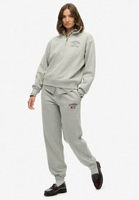 Vrouw in een lichtgrijze Superdry Athletics sweatshirt en bijpassende sweatpants, met bruine loafers, staand tegen een effen witte achtergrond.