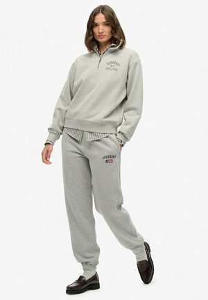 Vrouw in een lichtgrijze Superdry Athletics sweatshirt en bijpassende sweatpants, met bruine loafers, staand tegen een effen witte achtergrond.