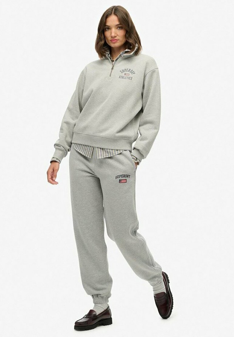 Vrouw in een lichtgrijze Superdry Athletics sweatshirt en bijpassende sweatpants, met bruine loafers, staand tegen een effen witte achtergrond.