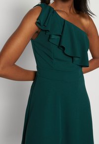 En axelklänning i mörkgrön tyg med ett volangaccent på axeln, figurnära bodice och flödande kjol. Slät textur, skräddarsydd design.