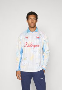 Maglietta da calcio a maniche lunghe in colori pastello, con un motivo floreale, dettagli blu e un logo rosso "KidSuper" sul davanti.