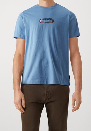 Homme portant un t-shirt bleu clair à manches courtes avec le texte "Hilfiger New York City 1985" et un pantalon marron.