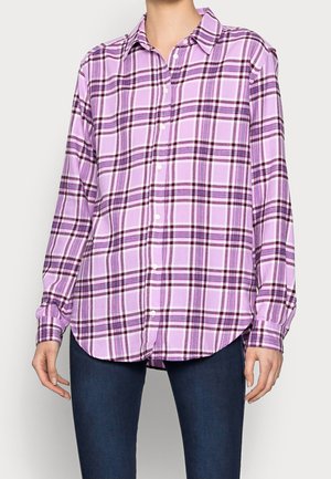 Kobieta ubrana w koszulę na guziki z długim rękawem w fioletowo-czarną kratę, zestawioną z ciemnoniebieskimi dżinsami.