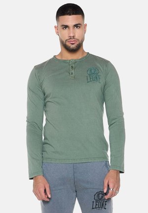Mann mit kurzen Haaren und Bart, der ein grünes langärmliges Henley-Shirt und graue Hosen mit „LEONE 1947“-Logos trägt, stehend vor weißem Hintergrund.