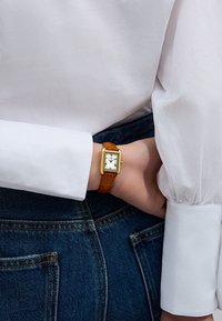 Montre rectangulaire dorée avec un cadran blanc et des chiffres romains noirs, associée à un bracelet en cuir marron, portée au poignet contre un denim.
