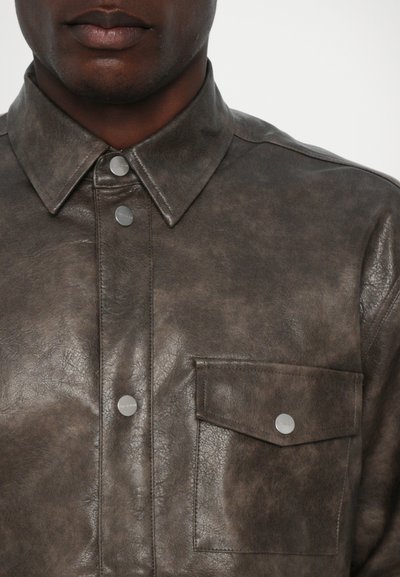 Chemise en cuir brun discret, avec un col pointu, des boutons-pression en argent et une poche poitrine à rabat. Fini texturé sur l'ensemble.