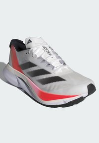 Zapatilla de running blanca con acentos en rojo y gris, parte superior de malla ligera, acabado texturizado, tres franjas negras prominentes, suela acolchada.