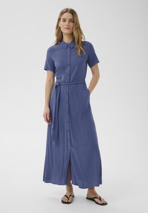 Młoda kobieta stoi z jedną dłonią w kieszeni, ubrana w niebieską sukienkę koszulową z guzikami, wiązaną w talii i czarne klapki.