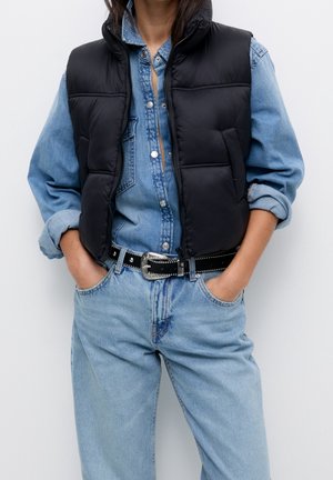 Person iført en sort dunvest over en lyseblå denimskjorte og matchende denimjeans med sort bælte, hænderne i lommerne.