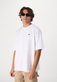 T-shirt bianca in cotone con maniche corte e collo rotondo. Presenta un piccolo logo nero sul lato sinistro del torace. Abbinata a pantaloni beige.