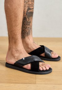 Sorte flip-flops med kryssede stropper, med en teksturert overflate og logo-detall. Glatt såle, brukt på bare føtter, med bakgrunn av tregulv.