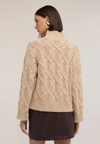 Beżowy sweter w warkoczyki z wysokim kołnierzem i ściągaczem, z misternymi warkoczowymi wzorami i długimi rękawami, noszony na brązowej skórzanej spódnicy.