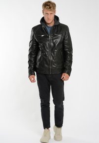 Svart läderjacka med dragkedja fram, sidfickor och en grå huva. Tillsammans med svartajeans och beige sneakers. Slät textur, avslappnad design.