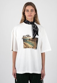 Νεαρή γυναίκα που φοράει λευκό φαρδύ T-shirt με την επιγραφή "Courtside Riviera Club", μαύρο κασκόλ με σχέδια στο λαιμό, κρίκους στα αυτιά και σκούρο παντελόνι.