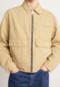 Jasnobeżowa kurtka z płótna z zamek błyskawicznym i dwoma przednimi kieszeniami, z kieszenią na piersi oraz metką marki na dolnej kieszeni.