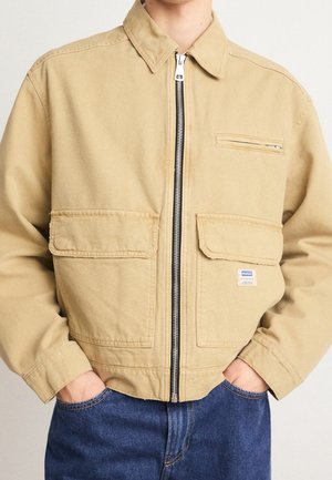 Chaqueta de lona color beige con cierre de cremallera y dos bolsillos frontales, que presenta un bolsillo en el pecho y una etiqueta de la marca en el bolsillo inferior.