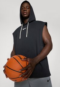Felpa nera senza maniche con cappuccio e cordini bianchi, logo Nike, abbinata a pantaloni grigi, mentre tiene in mano un pallone da basket arancione.