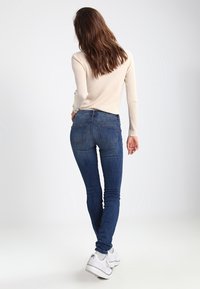 Top de manga larga de canalé en beige claro combinado con jeans ajustados azul oscuro y zapatillas blancas. Costuras visibles y detalles en el bolsillo trasero.